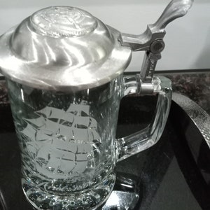 May include: A clear glass beer stein with a pewter lid and a hinged handle. The stein features an etched design of a sailing ship.