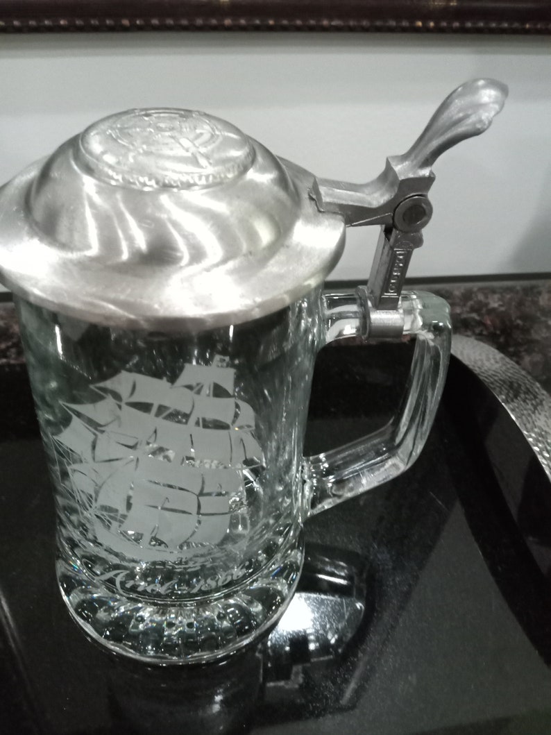 May include: A clear glass beer stein with a pewter lid and a hinged handle. The stein features an etched design of a sailing ship.