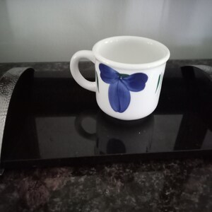 May include: White ceramic mug with a blue and green floral design. The mug is sitting on a black tray.