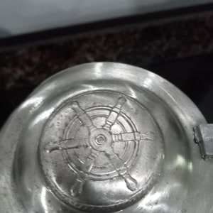 May include: A silver lid with a detailed embossed design of a ship's wheel.