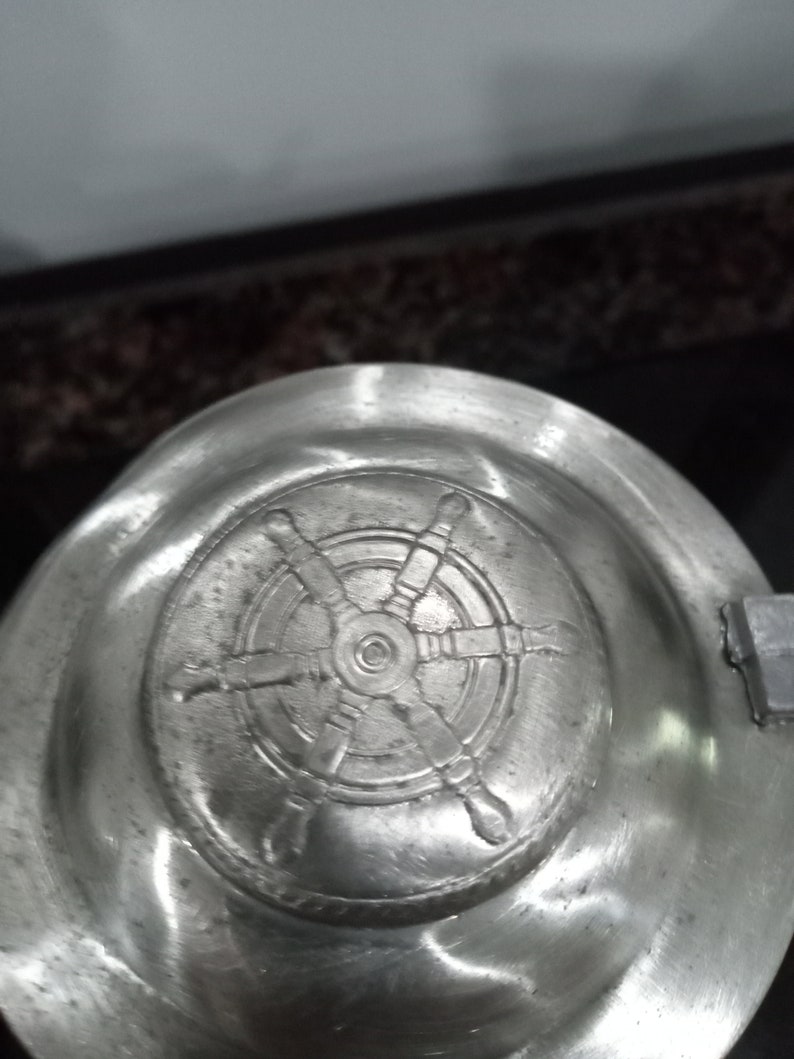 May include: A silver lid with a detailed embossed design of a ship's wheel.