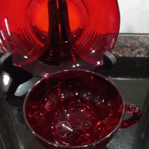 May include: A set of two ruby red glass teacups and saucers. The cups and saucers are both a deep red color and have a shiny finish.