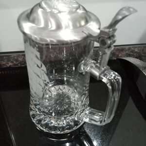 May include: A clear glass beer stein with a silver lid and a hinged handle. The stein has a decorative etched design on the front.