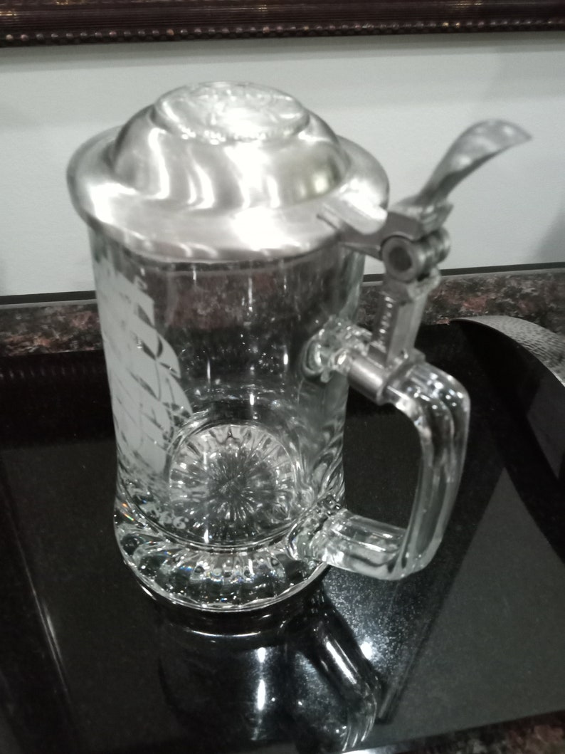 May include: A clear glass beer stein with a silver lid and a hinged handle. The stein has a decorative etched design on the front.