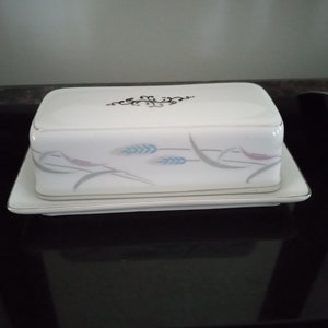 May include: White ceramic butter dish with a lid. The lid has a black floral design and the dish has a floral design with wheat stalks in gray and blue.