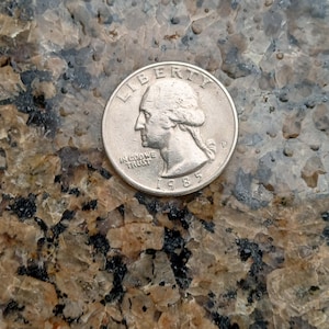 Może przedstawiać: Srebrna amerykańska moneta o wartości ćwierć dolara z napisem "Liberty" i "In God We Trust" oraz datą "1985".