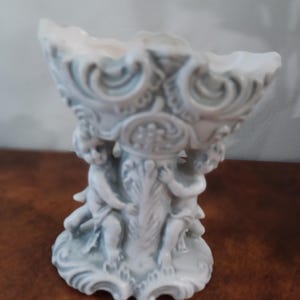 May include: A decorative white and grey bowl with an intricate design. The bowl is supported by three cherubic figures and features ornate scrollwork. The bowl is likely made of ceramic or porcelain and is suitable for decorative purposes.