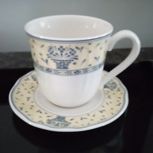 Puede incluir: Una taza de café de cerámica blanca con un patrón floral azul y amarillo. La taza está sentada sobre un platillo a juego.