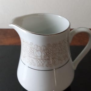 May include: White ceramic creamer with a silver rim and a floral design in shades of gray. The creamer has a handle and a rounded body.