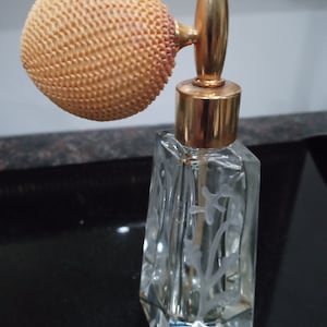 Può includere: Un flacone di profumo vintage in vetro con un tappo dorato e un atomizzatore rotondo testurizzato di colore arancio chiaro. Il flacone presenta un design floreale inciso.