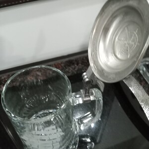 May include: A clear glass beer stein with a silver lid. The lid has a decorative design on the top.