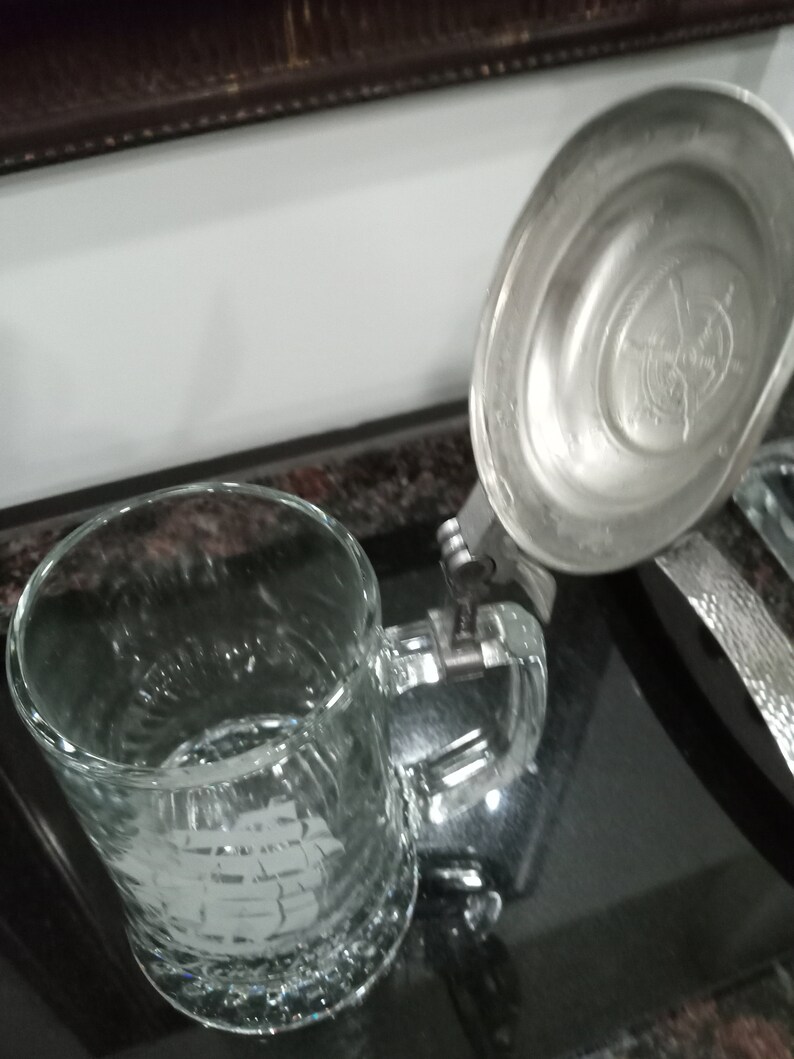 May include: A clear glass beer stein with a silver lid. The lid has a decorative design on the top.