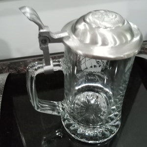 May include: A clear glass beer stein with a silver lid and a hinged handle. The lid has a raised design and the stein has a decorative etched design.