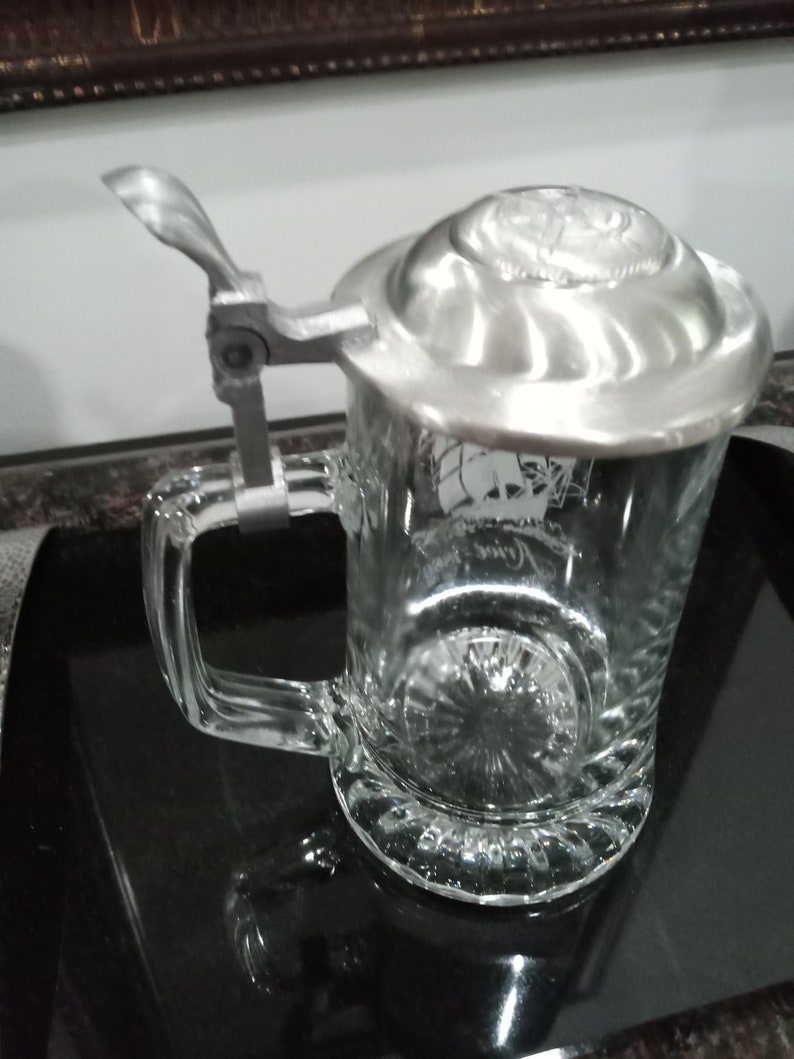 May include: A clear glass beer stein with a silver lid and a hinged handle. The lid has a raised design and the stein has a decorative etched design.