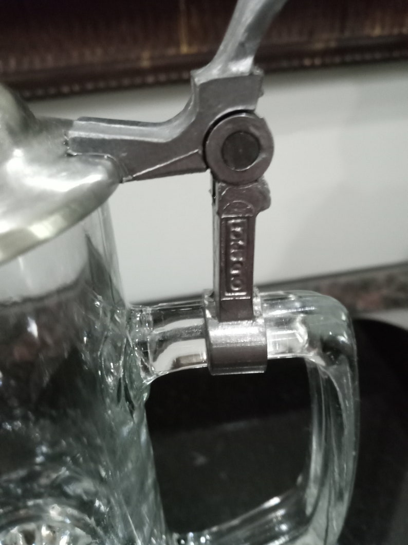 May include: Close-up of a silver and glass beer stein with a hinged lid and a handle. The lid has a decorative design and the handle is made of metal. The stein is made of clear glass with a textured surface.