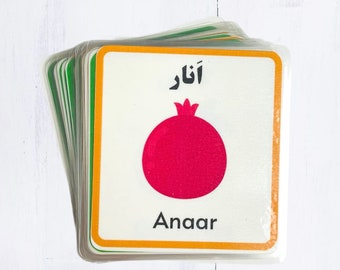 Pode incluir: Um cartão branco com uma ilustração de romã vermelha e as palavras "Anar" em inglês e árabe. O cartão é rodeado por uma borda amarela e uma borda verde.