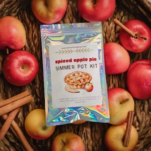 May include: A silver holographic pouch with the text "spiced apple pie SIMMER POT KIT" and a drawing of an apple pie. The pouch is surrounded by red apples and cinnamon sticks on a woven basket.
