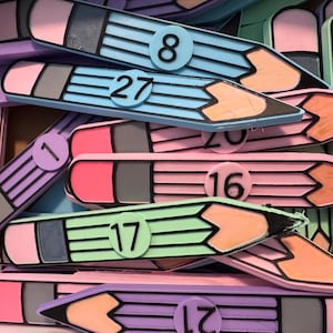 Puede incluir: Una colección de recortes planos con forma de lápiz en tonos pastel azul, rosa, morado y verde. Cada lápiz tiene una goma negra, una virola gris y una etiqueta circular con un número impreso en negro, incluyendo 1, 7, 8, 16, 17, 19, 20, 21 y 27.