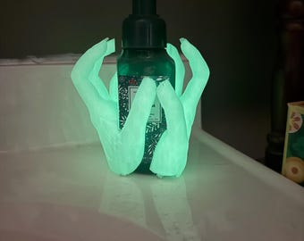 Puede incluir: Un dispensador de jabón de manos verde con una bomba negra, sostenido por un soporte en forma de mano verde brillante. El frasco de jabón tiene un diseño floral. El soporte está sobre una superficie blanca.
