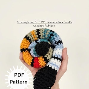 May include: A colorful crochet snake toy with a black head and button eyes. The body is a spiral of various colors, including yellow, orange, blue, and white. The text "Birmingham, AL 1995 Temperature Snake Crochet Pattern" is above the snake. A "PDF Pattern" logo is in the lower left.