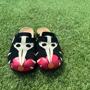 May include: A pair of black suede clogs with a white and red floral design. The clogs have a skull and beak design on the front.
