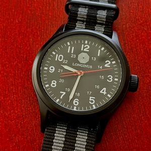 May include: A black wristwatch with a black face and white numbers. The watch has a black and gray striped nylon strap. The watch face has the word "LONGINUS" printed on it.