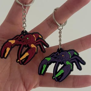 May include: Two lobster-shaped keychains. One is red with yellow accents, the other is purple with green accents. Both have black outlines and silver key rings. The keychains are held up against a light background.
