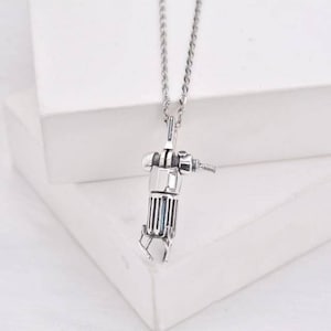 Puede incluir: Un collar de plata con un colgante detallado con forma de robot. El robot tiene un cuerpo cilíndrico con engranajes y una broca.