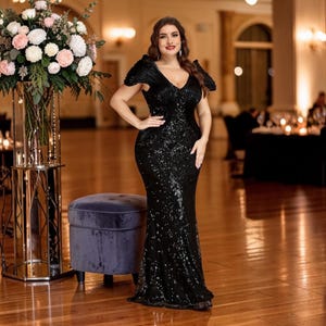 K&ouml;nnte beinhalten: Ein schwarzes Pailletten-Abendkleid mit V-Ausschnitt und kurzen &Auml;rmeln. Das Kleid hat eine figurbetonte Silhouette, die sich am Saum ausstellt. Die Frau steht in einem Ballsaal mit einem Blumengesteck und einem Samt-Hocker.