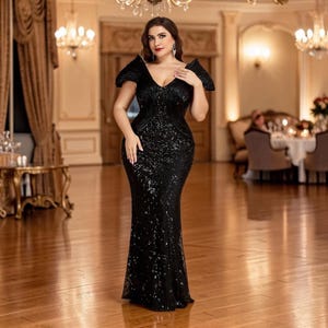 K&ouml;nnte beinhalten: Ein schwarzes Abendkleid mit Pailletten, V-Ausschnitt und strukturierten Schultern. Das bodenlange Kleid hat ein figurbetontes Oberteil und einen ausgestellten Rock. Die Frau steht in einem gut beleuchteten Raum mit Kronleuchtern und aufwendiger Dekoration.