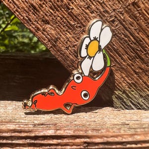 May include: A red enamel pin featuring a cartoon character with a long body, two large white eyes, and a small mouth. A large white flower with a yellow centre and a green stem is attached to the top of the character's head, outlined in gold metal.