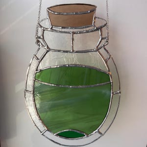 Puede incluir: Botella de poción de vidriera con líquido verde en el interior y un tapón de corcho. La botella está delineada en plata y cuelga de una cadena. El vidrio tiene una apariencia texturizada, y el líquido verde es visible a través del vidrio transparente.