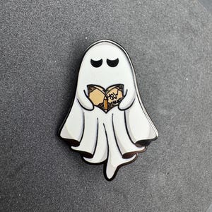 以下が含まれることがあります： 黒い目と黒いアウトラインの白い幽霊型のエナメルピン。幽霊は表紙に「How to Haunt」という言葉が書かれた本を持っています。ピンは灰色の表面にあります。