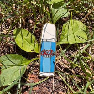 Peut inclure: Une broche en émail métallique en forme de thermos vintage avec un bouchon blanc et un corps bleu clair. Le centre du thermos présente un logo ovale rouge avec le texte OhD en écriture blanche et le mot Diner en plus petites lettres blanches.