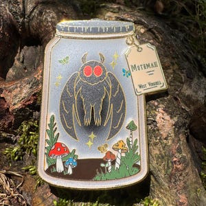 May include: A gold-rimmed enamel pin shaped like a mason jar containing a black Mothman figure with glowing red eyes. The jar features colorful mushrooms, green foliage, and a hanging tag labeled Mothman West Virginia with small star and butterfly accents.
