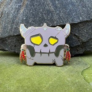 May include: A gray and gold enamel pin featuring a cartoon skull with yellow eyes and red feathers on the sides.