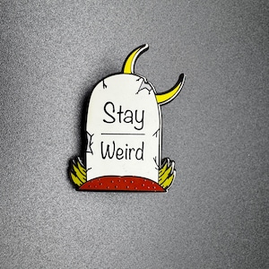 May include: A white enamel pin shaped like a tombstone with a yellow crescent moon on top. The tombstone says "Stay Weird" in black lettering.
