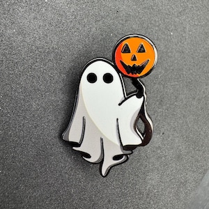 以下が含まれることがあります： ハロウィーンをテーマにしたエナメルピン。黒い目と銀色の輪郭を持つ白い幽霊が、オレンジ色のジャック・オー・ランタンを持っています。ジャック・オー・ランタンは黒い顔と黒い茎をしています。ピンは灰色の背景に配置されています。