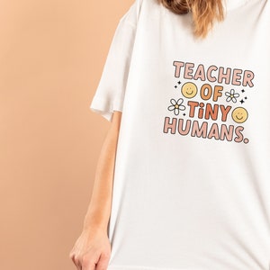 May include: White t-shirt with the text "TEACHER OF TINY HUMANS." in pink and orange lettering with a daisy and smiley face graphic.