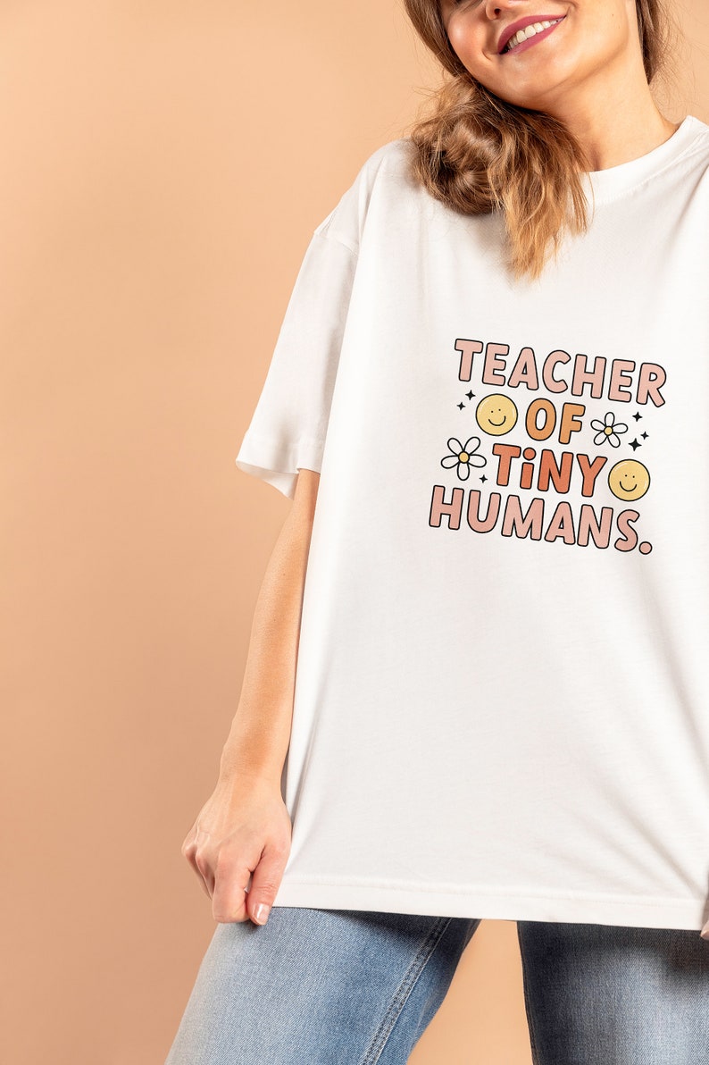 May include: White t-shirt with the text "TEACHER OF TINY HUMANS." in pink and orange lettering with a daisy and smiley face graphic.