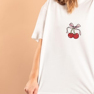 May include: White t-shirt with a cartoon cherry design. The graphic features two red cherries and a pink bow. The shirt has a relaxed, oversized fit, ideal for casual wear.