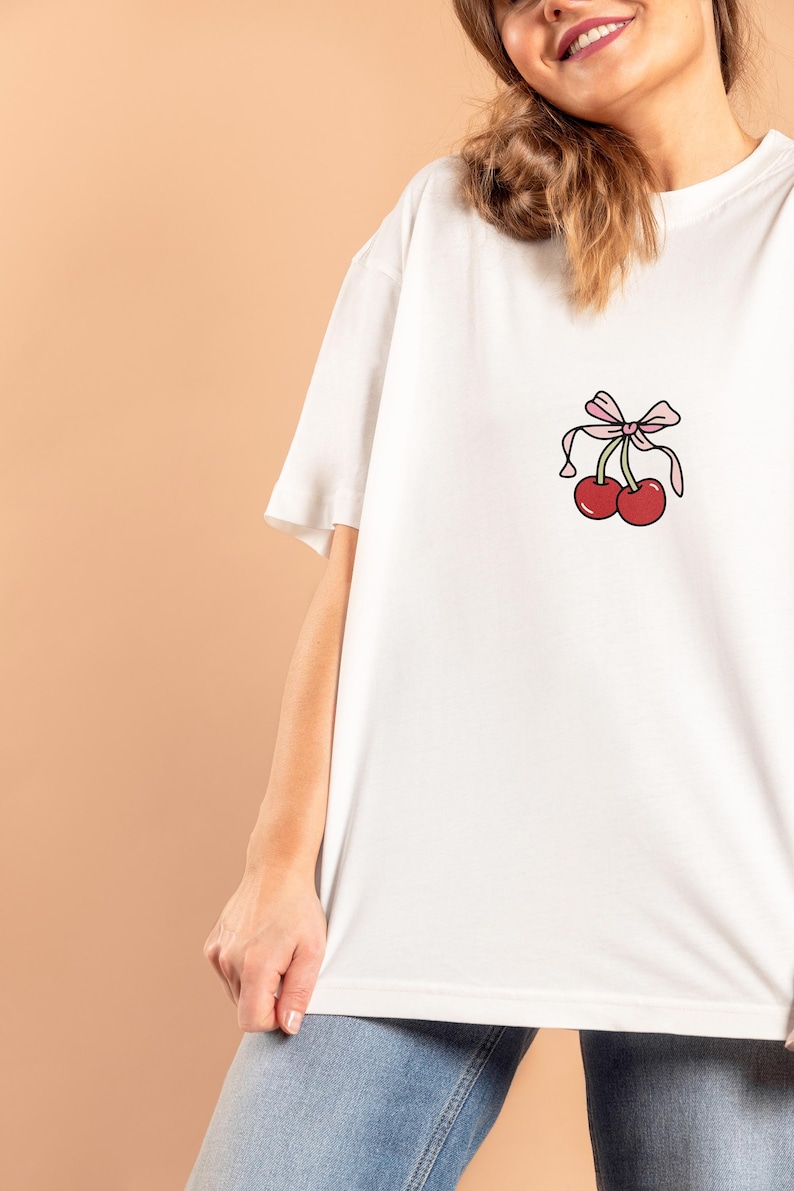 May include: White t-shirt with a cartoon cherry design. The graphic features two red cherries and a pink bow. The shirt has a relaxed, oversized fit, ideal for casual wear.