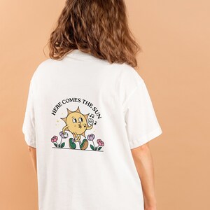 May include: White t-shirt featuring a cartoon sun graphic and the text "HERE COMES THE SUN". The sun character is yellow with green shoes and is holding a musical note. The design includes flowers.