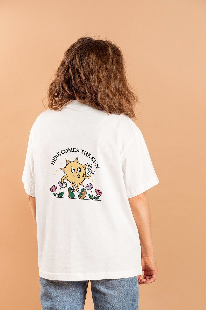 May include: White t-shirt featuring a cartoon sun graphic and the text "HERE COMES THE SUN". The sun character is yellow with green shoes and is holding a musical note. The design includes flowers.