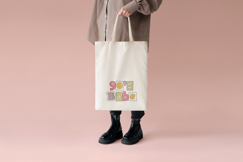 May include: A white canvas tote bag with the text "90's Babe" in colorful block letters. The letters are decorated with smiley faces and stars.