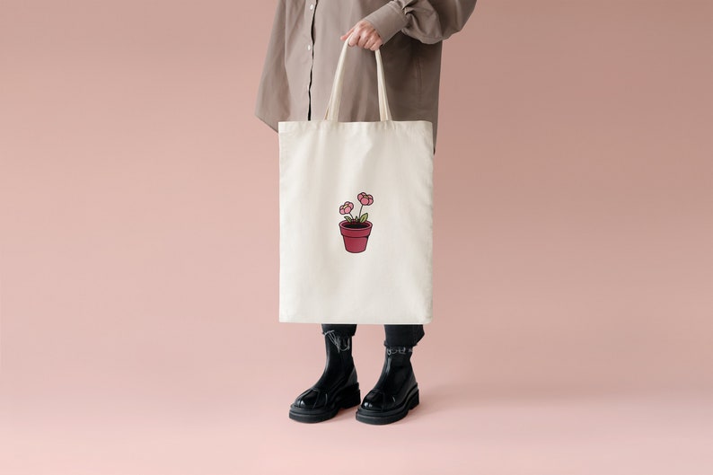 May include: A white tote bag with a pink flower pot design. The bag has brown straps and is being held by a person.