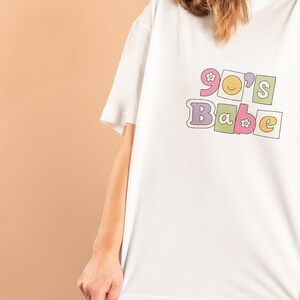 May include: White t-shirt with a graphic print of "90's Babe" in colorful block letters with flowers and a smiley face.