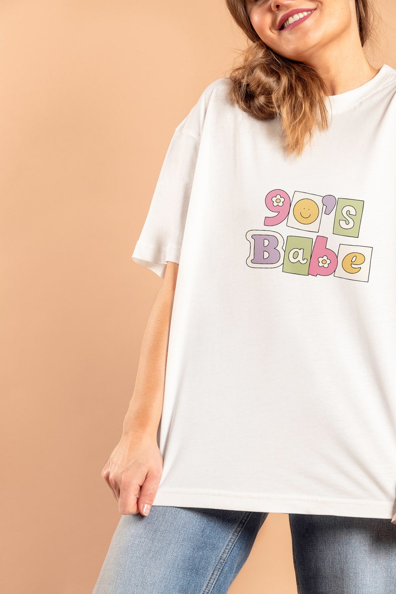 May include: White t-shirt with a graphic print of "90's Babe" in colorful block letters with flowers and a smiley face.