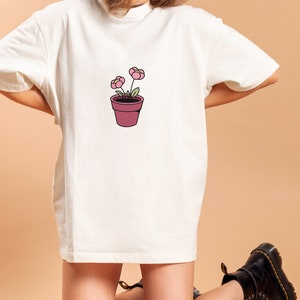 May include: A white oversized t-shirt with a simple illustration of a potted plant with two pink flowers. The person wearing the shirt is kneeling on a light brown surface.