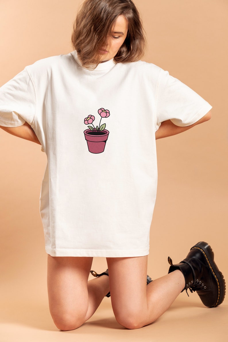 May include: A white oversized t-shirt with a simple illustration of a potted plant with two pink flowers. The person wearing the shirt is kneeling on a light brown surface.